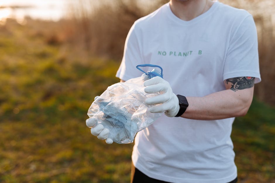 A person wearing a white T-shirt with the slogan 'No Plant B' and white gloves is holding a clear plastic bag filled with various discarded items including crumpled paper and small debris. The individual appears to be outdoors on a natural terrain with blurred greenery and brownish foliage in the background, illuminated by natural sunlight. The person also has a tattoo on their left arm and is wearing a black wristwatch. The scene suggests a context of environmental cleanup or waste collection, aligning with practices of independent rubbish removal services, although no specific equipment such as trucks or tools are visible. The image captures a moment of careful handling of waste in an open setting, emphasizing environmentally conscious waste management and the importance of proper rubbish disposal or recycling efforts.