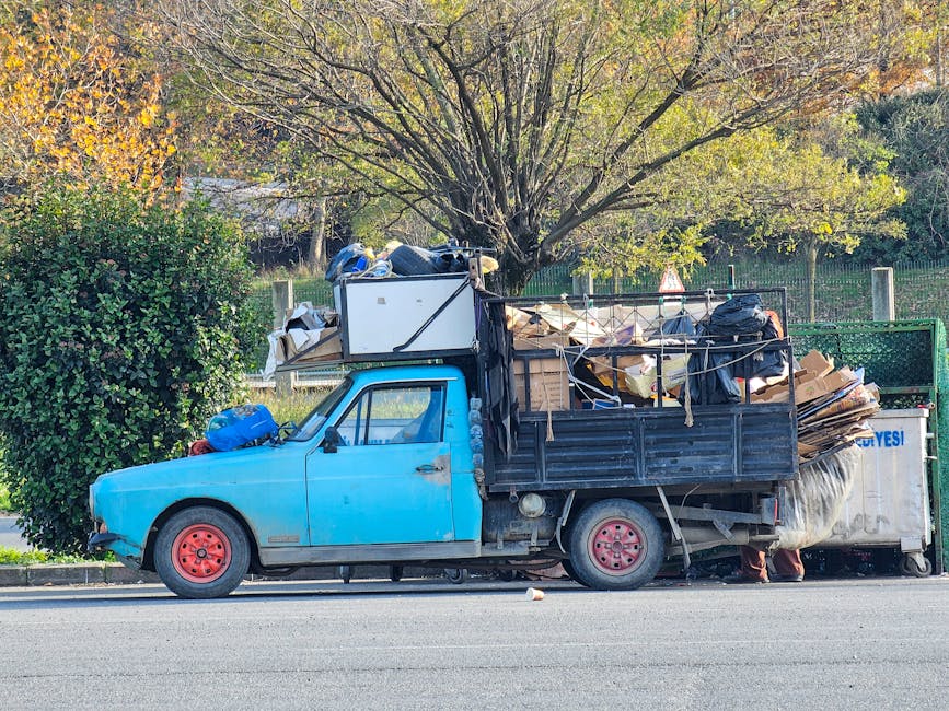 A small, older model light blue flatbed truck parked on the side of a street with a black metal railing behind it. The truck is loaded with various types of rubbish, including cardboard boxes, black plastic garbage bags, and miscellaneous debris, which are stacked haphazardly and secured with ropes. The cargo extends above the truck's cab and onto the flatbed, filling the vehicle's entire bed area. The truck's wheels are red and show some signs of wear. In the background, there is a large tree with a mix of green and yellow leaves indicating autumn, and a green wooded area enclosed by a fence. The scene is illuminated by natural daylight, highlighting the textures and colors of the rubbish and the environment. This image exemplifies a private rubbish collection that might be handled by waste removal specialists such as Rubbish Removal Wandsworth, providing an alternative to municipal waste disposal services.