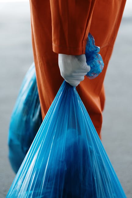 Close-up of a person wearing protective white gloves and bright orange worker's overalls, holding a tightly tied blue plastic rubbish bag by its gathered top. The person is standing outdoors on a paved surface, with a blurred background suggesting an open area such as a street or driveway. The plastic bag appears full, with the material stretched and semi-transparent, revealing the contents inside. The scene emphasizes proper handling and collection of waste materials, consistent with private removal or alternative waste disposal services provided by companies like Rubbish Removal Wandsworth. The lighting is natural and even, highlighting the textures of the gloves, overalls, and the smooth surface of the plastic bag. This image captures the typical act of waste collection from a waste management professional engaged in rubbish removal activities, reflecting the inspection and handling processes involved in onsite clearance or independent rubbish disposal routines.